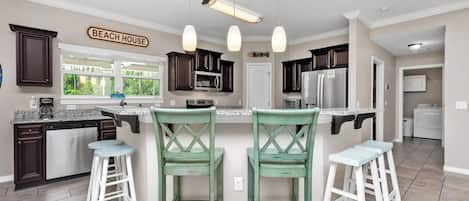 A kitchen with dark wood cabinets, a central island with stools, pendant lighting, and a sign reading 'BEACH HOUSE'.