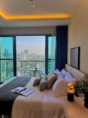 A hotel room with a large bed, a bedside table with a lamp, a view of a cityscape through large windows, and a small balcony table. - 5 min Metro| KingBed | Kitchen full | Netflix WiFi (Watthana)
