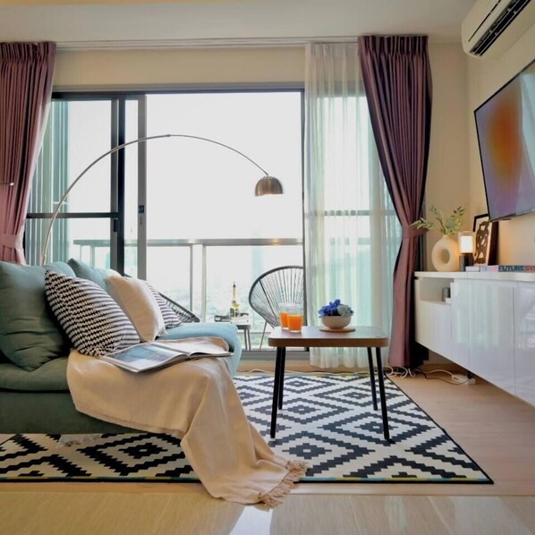 A modern living room with a sofa, a coffee table, a lamp, and a TV. - 5 min Metro| KingBed | Kitchen full | Netflix WiFi (Watthana)