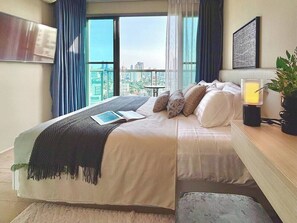 Ein modernes Hotelzimmer mit einem großen Bett, einem Nachttisch mit Lampe, Blick auf die Stadt durch große Fenster und einem angebrachten Fernseher.