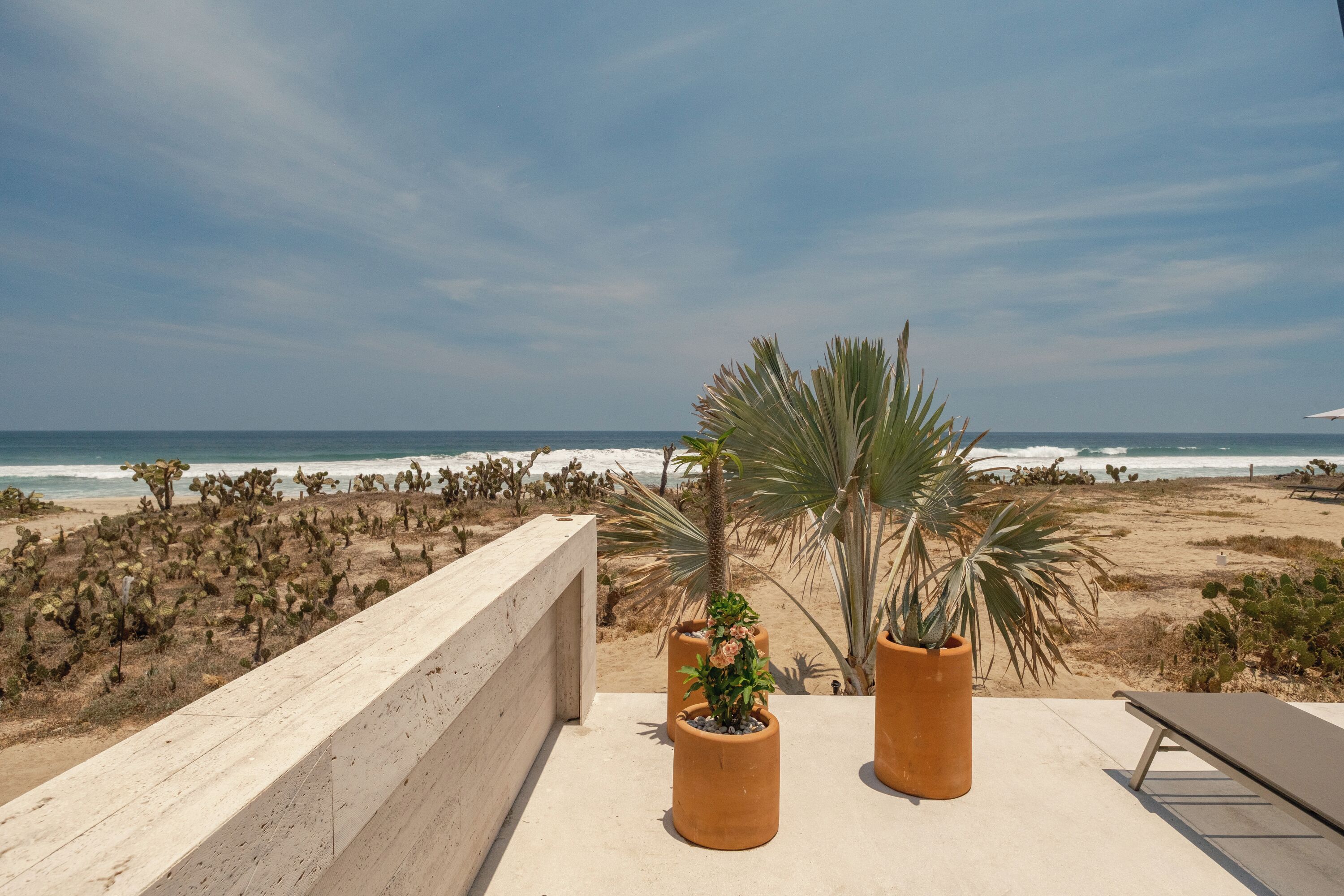A balcony with a view of the beach and ocean, potted plants, and a lounge chair.