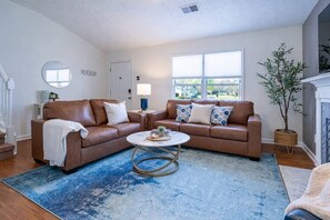 Ein Wohnzimmer mit zwei braunen Ledersofas, einem runden Couchtisch, einem blauen Teppich, einer Topfpflanze und einem Kamin.