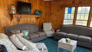 A cozy living room with wooden walls, a television, two sofas, a coffee table, and a large window. - Lakefront cottage #1 with private beach on Lake Huron, AC, shared Hot Tub & Pool (Lexington)