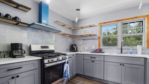 A modern kitchen with grey cabinets, a stove, a coffee maker, and a window with a view of trees. - Lakefront cottage #1 with private beach on Lake Huron, AC, shared Hot Tub & Pool (Lexington)