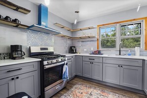 A modern kitchen with grey cabinets, a stove, a coffee maker, and a window with a view of trees.