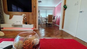 Un salon avec un téléviseur, un saladier en verre contenant des objets décoratifs, un tapis rouge et, au fond, un coin repas.