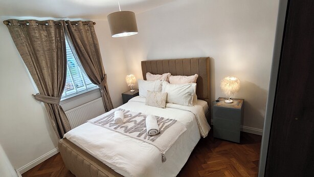 Une chambre à coucher comprenant un lit, deux tables de chevet, une fenêtre avec des rideaux, un luminaire suspendu et un radiateur.