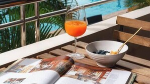 Escena junto a la piscina con un libro, un vaso de zumo de naranja y un cuenco de fruta sobre una mesa de madera.
