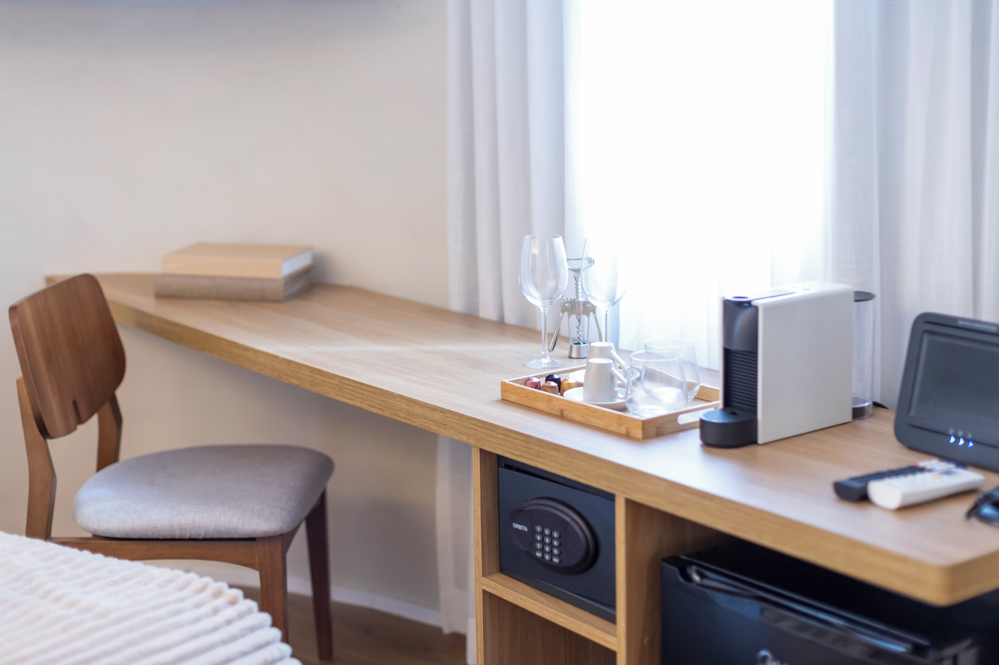 A hotel room with a wooden desk, a chair, a bedside table, a bedside lamp, a coffee maker, a remote control, and a window with curtains.