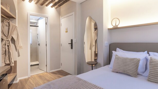 A hotel room with a bed, a bedside table, a mirror, a robe hanging on a rack, and a door leading to a bathroom. - Stay Boutique Ibirapuera (São Paulo)