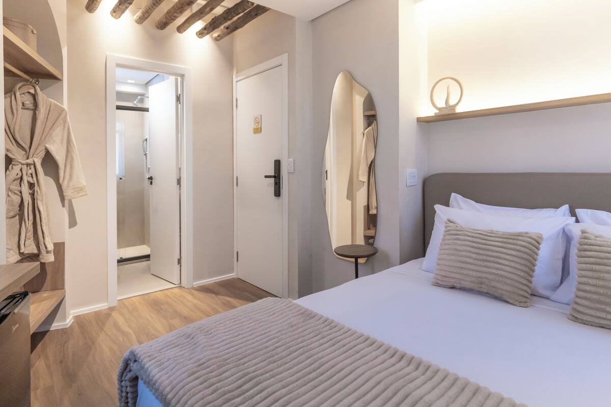 Quarto de hotel com uma cama, mesa de cabeceira, espelho, roupão pendurado em um suporte e porta para um banheiro.