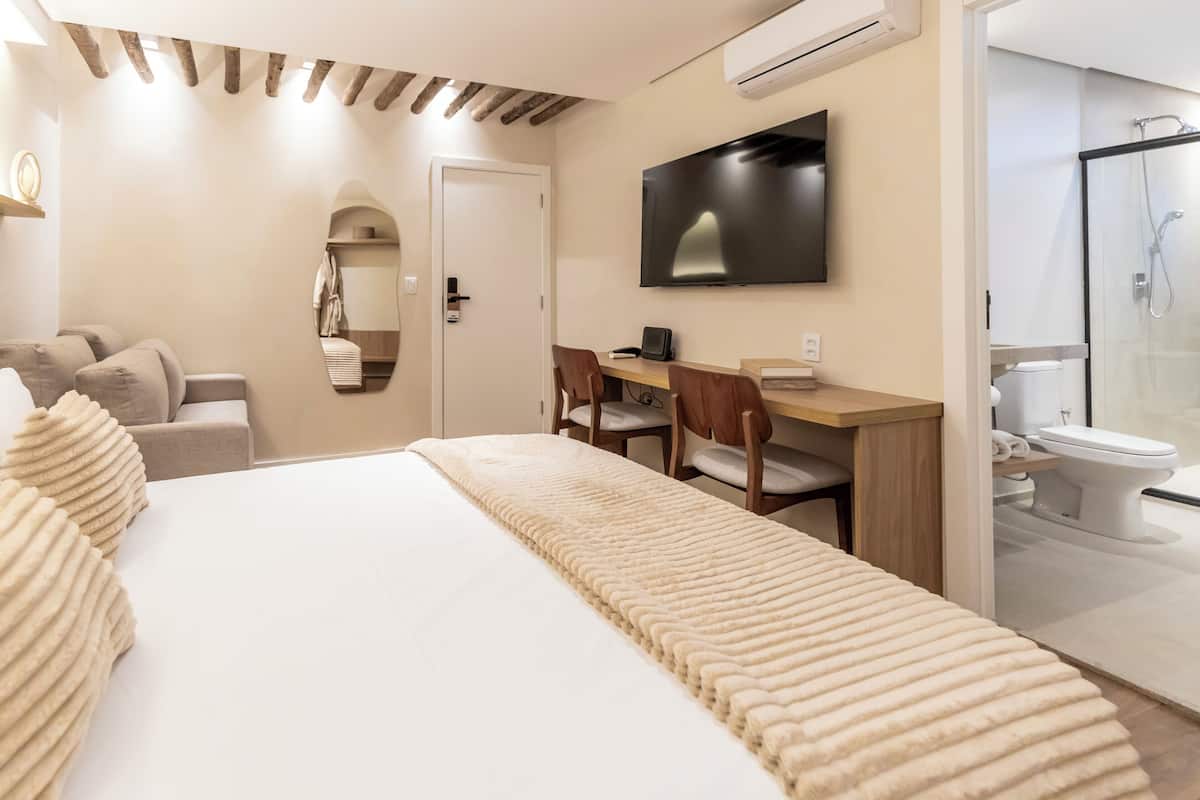 Quarto de hotel com cama, secretária, cadeira, televisão, chuveiro e sanita.