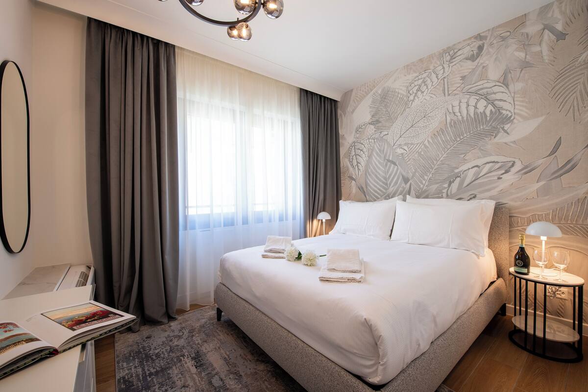 A hotel room with a large bed, a bedside table with a bottle and glasses, a desk with a lamp, and a wall with a leaf pattern.