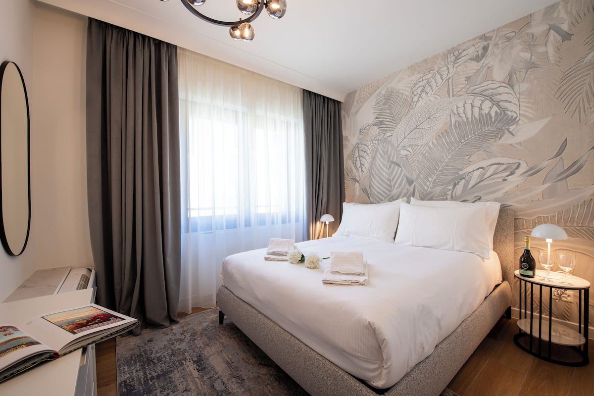 A hotel room with a large bed, a bedside table with a bottle and glasses, a desk with a lamp, and a wall with a leaf pattern.