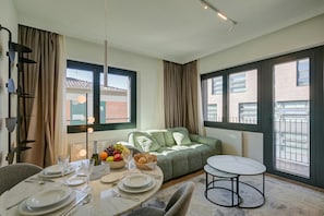 A modern living room with a sofa, a dining table set, and large windows with a view of buildings.