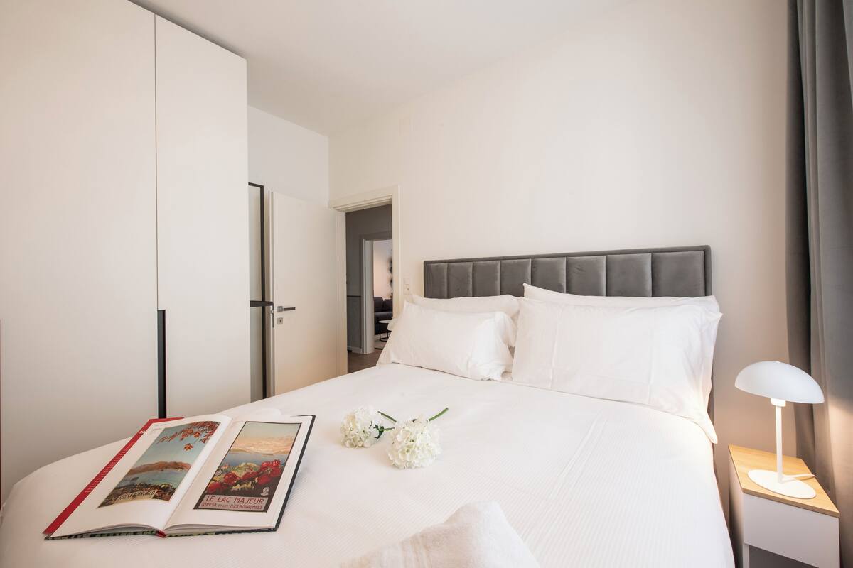 A hotel room with a bed, a nightstand with a lamp, a wardrobe, and an open book on the bed.
