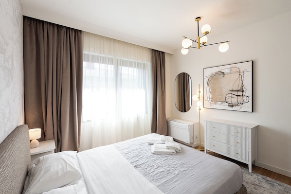A modern bedroom with a bed, curtains, a radiator, a bedside table, a lamp, and a wall-mounted light fixture.