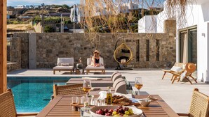 A poolside dining area with a table set for two, a wicker chair, and a hanging chair. - Vendema Residence Mykonos (Mikonos)