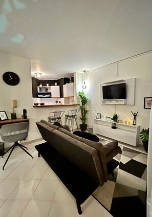 Un salon moderne avec un canapé, des fauteuils, un téléviseur et un coin cuisine à l’arrière-plan.