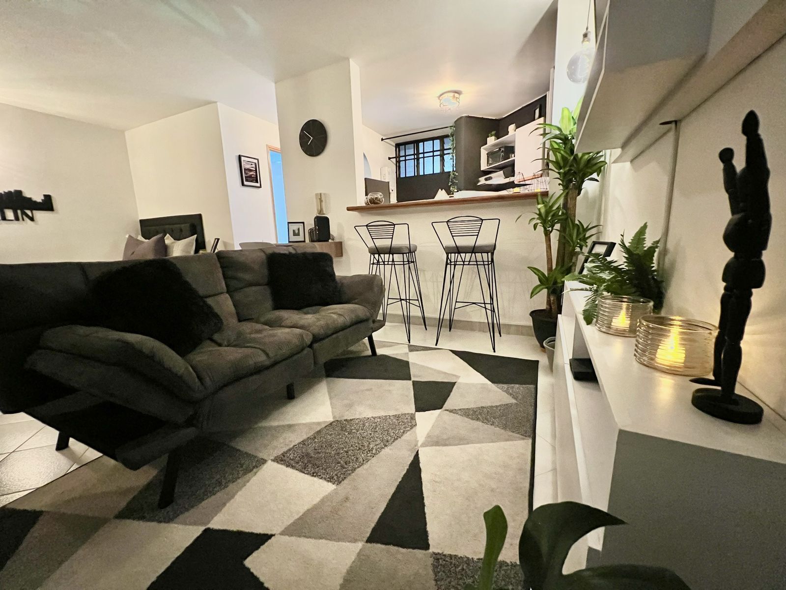 Un salon moderne avec un canapé, un tapis à motifs géométriques, un coin repas avec des chaises et une cuisine en arrière-plan.