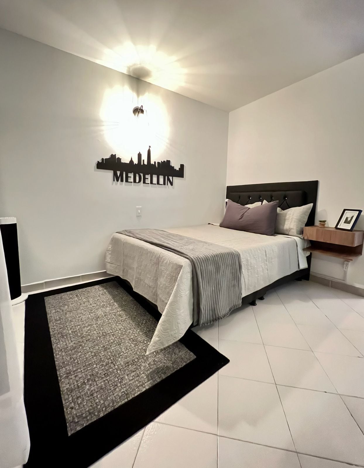 Une chambre à coucher comprenant un lit, une table de chevet, une lampe fixée au mur et un autocollant représentant un paysage urbain.
