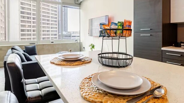 Una cocina moderna con una mesa de comedor para dos, una cesta de metal negro con aperitivos, y vistas a la ciudad a través de grandes ventanales.