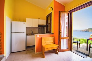 A compact kitchen with a refrigerator, stove, and cabinets, a wooden bench, and a view of the sea through an open door. - Comfortable House with Large Terrace Sea View - Pistis - Green Coast-Arbus (Sardegna)