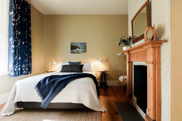 A bedroom with a bed, bedside lamps, a painting, a fireplace, and a window with curtains.