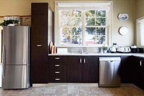 A modern kitchen with a stainless steel refrigerator, dark cabinetry, a dishwasher, and a window with a view of trees.