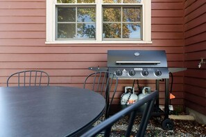 A grill with a gas tank, a table, and chairs outside a house with a window.