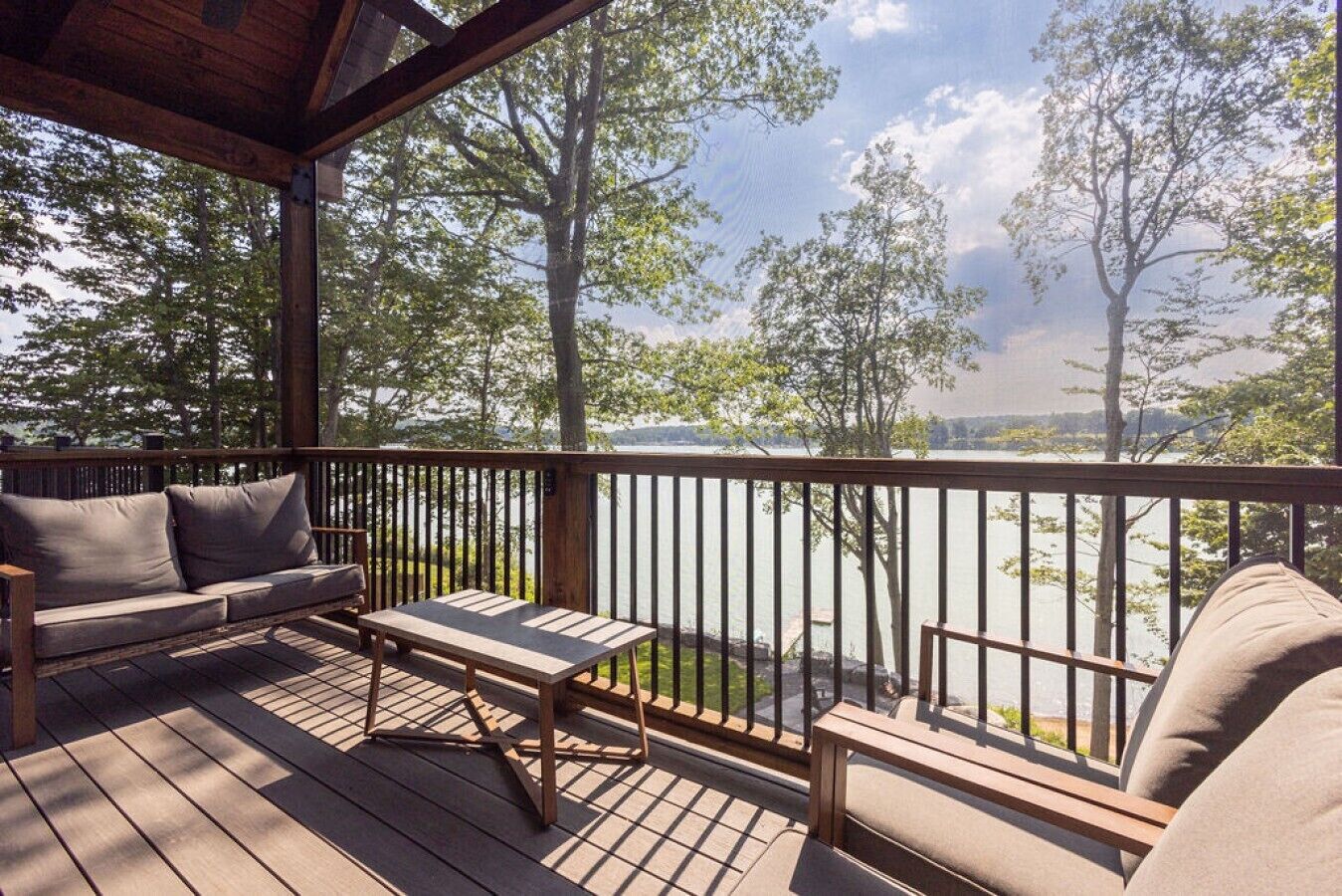 Terraza de madera con un sofá y una mesa, con vista a un cuerpo de agua y árboles.