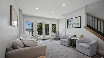 A spacious living room with a grey sofa, two armchairs, a small table, and a staircase.