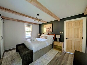 Un dormitorio con cama, mesitas de noche, un banco, una ventana, una puerta y un ventilador de techo.