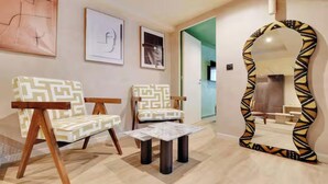 Um quarto com duas cadeiras estampadas, uma mesinha, um espelho com moldura geométrica e obras de arte nas paredes.