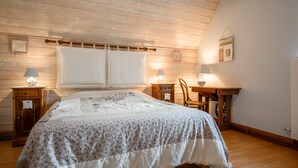 A bedroom with a wooden ceiling, a bed with a white and floral bedspread, two bedside tables with lamps, and a small wooden chair. - Guest House 'Suite Familiale - Kobo-Maya' with Mountain View and Shared Garden (Orbey)