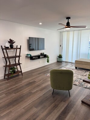 Ein modernes Wohnzimmer mit Flachbildfernseher, Holzboden, Deckenventilator und einem grünen Sessel.
