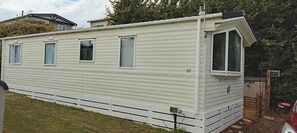 A single-story mobile home with a front porch, a small attached garage, and a fenced yard. - Stay 6 - Pool - Parking - Beachside - Pet Friendly (Norwich)