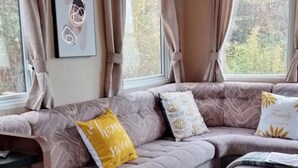 A cozy RV interior with a sectional sofa, a dining table, and large windows with curtains. - Stay 6 - Pool - Parking - Beachside - Pet Friendly (Norwich)