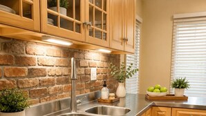 Une cuisine dotée d’un évier en acier inoxydable, de meubles de rangement en bois, d’un mur en briques et d’une fenêtre avec des stores.