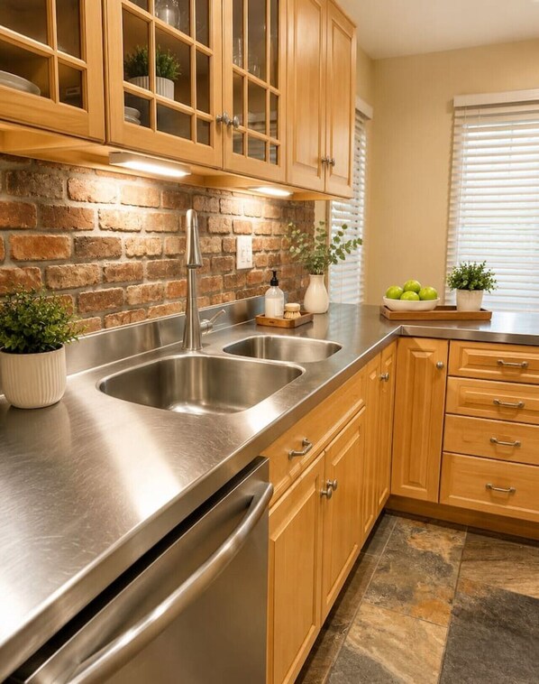 A kitchen with a stainless steel sink, wooden cabinets, a brick wall, and a window with blinds.