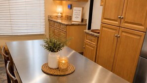 Une cuisine moderne avec une table en acier inoxydable, des chaises en bois, un set de table en fibres naturelles, une plante en pot et une bougie allumée.