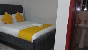 Una cama con cabecera oscura, ropa de cama blanca y amarilla, y una toalla enrollada.
