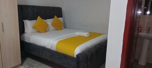Ein Bett mit dunklem Kopfteil, weißer und gelber Bettwäsche sowie einem gerollten Handtuch.