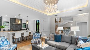 Ein modernes Wohnzimmer mit einer grauen Couch, gemusterten Sesseln und einem Essbereich mit weißen Schränken und einer schwarzen Spritzschutzleiste.