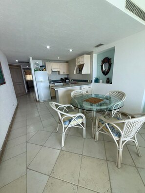 A modern kitchen with white cabinets, a refrigerator, and a dining area with a glass table and wicker chairs. - Oceanfront Condo at Bay View Grand | Infinity Pool, Gym, and Tennis Court (Puerto Vallarta)