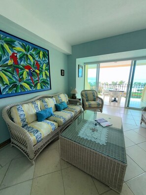 A living room with a wicker sofa, a glass coffee table, and a chair. There is a painting on the wall and a view of the outdoors through a glass door. - Oceanfront Condo at Bay View Grand | Infinity Pool, Gym, and Tennis Court (Puerto Vallarta)