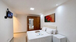 A bedroom with a bed, bedside table, a small refrigerator, a TV mounted on the wall, and a painting on the wall. - Suíte com Jacuzzi Vista de Escarpas 1 (Capitólio)