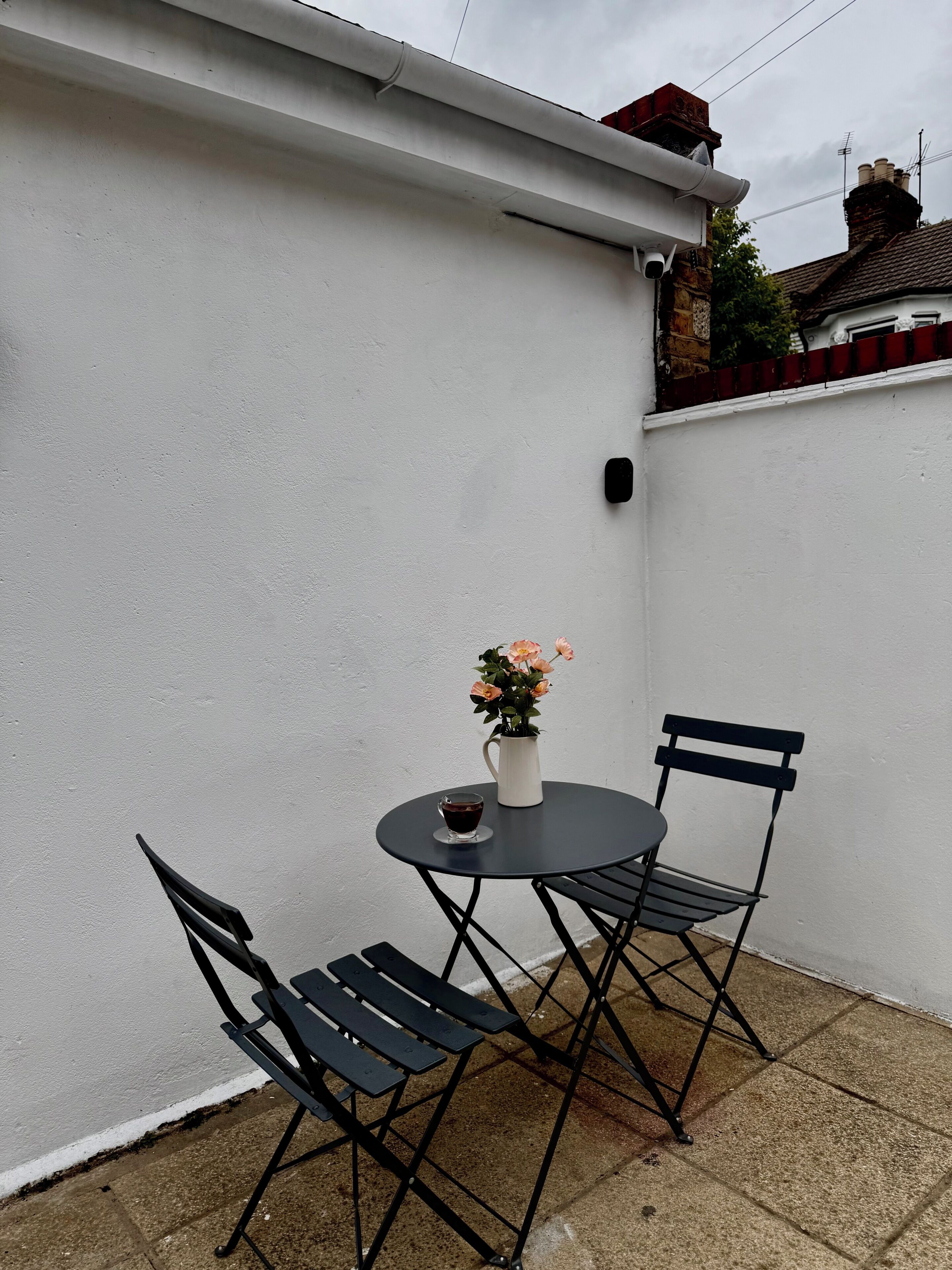 Une terrasse avec une table ronde, deux chaises, un vase de fleurs et un appareil fixé au mur.