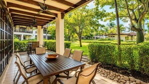 Une terrasse extérieure avec une table et des chaises en bois, un ventilateur de plafond et une vue sur un jardin bien entretenu et les bâtiments voisins.