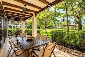 Eine Terrasse im Freien mit einem Holztisch und -stühlen, einem Deckenventilator und Blick auf einen gepflegten Garten sowie angrenzende Gebäude.